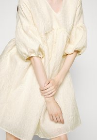 Personne portant une robe crème texturée avec des manches bouffantes, tenant un bras avec la main opposée devant son corps.
