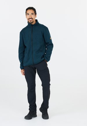 Vihreä fleece-takki korkean kauluksen, täyden vetoketjun, kahden sivutaskun ja tekstuuripinnan kanssa. Yhdistetty mustiin ulkoiluhousuihin ja saappaisiin.