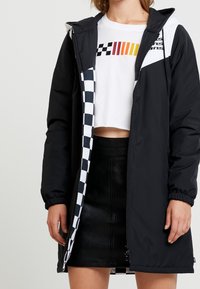 Person som bär en svart huvad jacka med rutigt foder, vit kort t-shirt med racingränder, och svart läder mini-kjol.