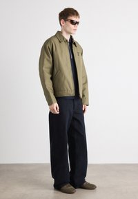 Veste olive avec col, fermeture éclair et poche zippée, associée à un pantalon large marine foncé et des chaussures marron. Le mannequin porte des lunettes de soleil.