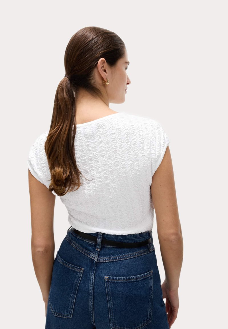 Cropped top bianco texturizzato con maniche a cappuccio, abbinato a jeans in denim scuro a vita alta. Presenta una cintura nera e un semplice design sul retro.