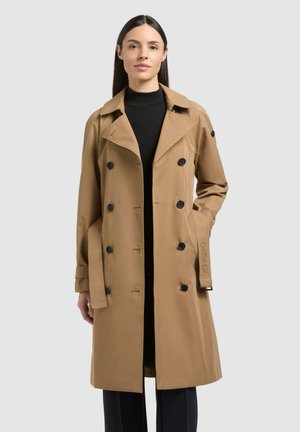 Frau steht und trägt einen beige-doppelreihigen Trenchcoat über einem schwarzen Rollkragenpullover und schwarzen Hosen vor einem schlichten hellen Hintergrund.