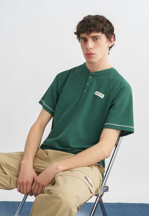 Młody mężczyzna z kręconymi włosami siedzący na metalowym krześle, ubrany w zieloną koszulkę typu henley z krótkim rękawem i beżowe spodnie cargo na jednolitym tle.