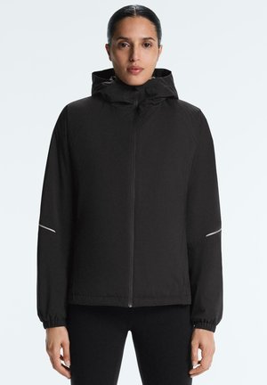 Kvinde med mørkt hår trukket tilbage, iført en sort hættetrøje med lynlås og elastiske ærmekanter, stående mod en ensfarvet lys baggrund.