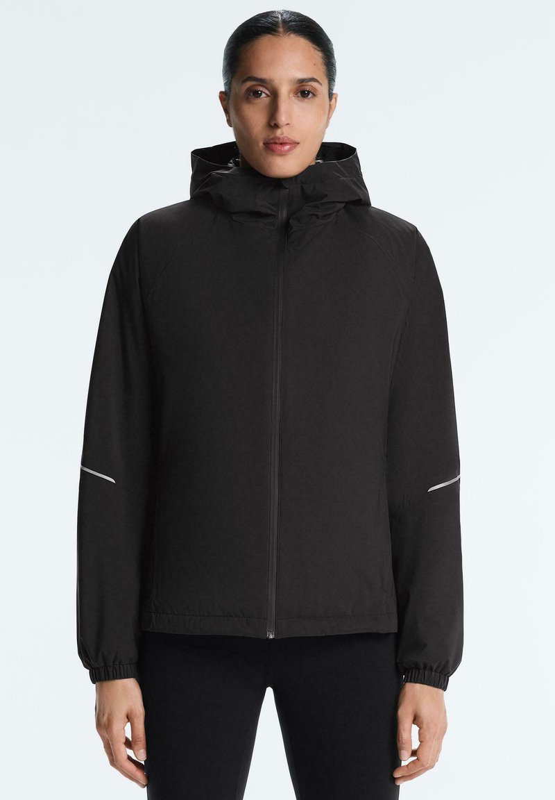 Kvinde med mørkt hår trukket tilbage, iført en sort hættetrøje med lynlås og elastiske ærmekanter, stående mod en ensfarvet lys baggrund.