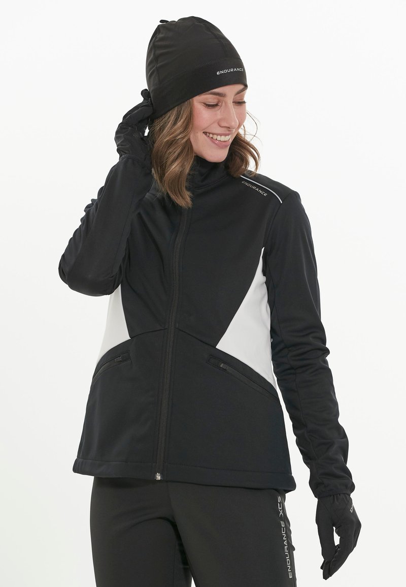 Black and white zip-up jacket with a high collar, featuring zippered pockets and a fitted design. Worn with a black beanie and gloves.
