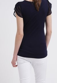 Mörkblå top med korta, genomskinliga ärmar; figurnära design; slät textur; kombinerad med vita byxor; sedd bakifrån.