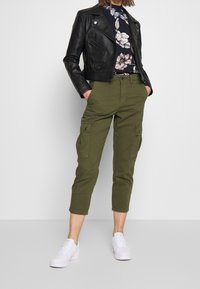 Schwarze Lederjacke in Cropped-Form, Blumenshirt, olivgrüne Cargo-Hosen und weiße Sneaker. Die Jacke verfügt über Reißverschlüsse; die Hosen haben Seitentaschen.