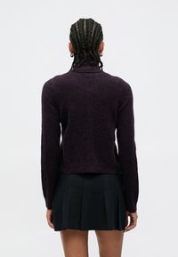 Vijoličen, oprijet pulover z visokim ovratnikom in dolgimi rokavi iz teksturirane pletene tkanine. Kombiniran s črno plisirano krilo, prikazan od zadaj.
