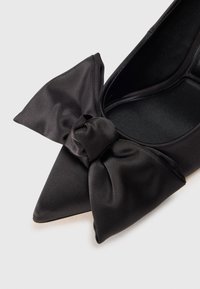 Chaussure plate noire à bout pointu avec un grand nœud en satin sur l’avant, présentée sur un fond clair.
