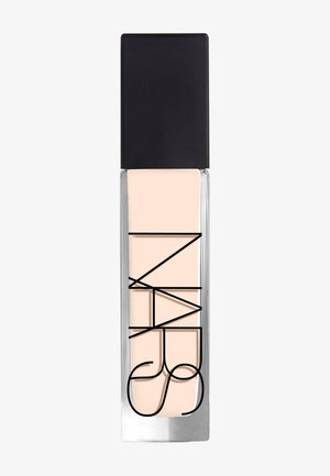 Rechteckige Glasflasche mit schwarzem Verschluss, die eine hellbeige flüssige Foundation enthält, mit fettem schwarzen Text auf der Vorderseite.