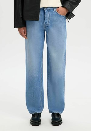 Persoon draagt lichtblauwe rechte jeans, zwarte schoenen, zwarte leren jas en een crèmekleurige trui met één hand in de zak.