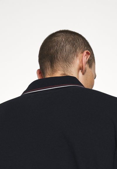 Navy polo shirt with a structured collar featuring three stripes in red and white. The fabric appears smooth, with a slightly textured finish.