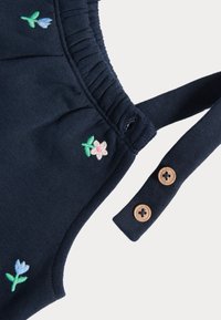 Marinblå plagg med blommönster i grönt, rosa och ljusblått. Har elastisk rynkning och träknappdetaljer.