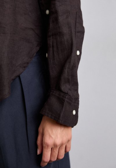 Chemise en lin marron foncé avec une coupe décontractée, présentant un poignet boutonné et des détails de couture visibles, assortie à un bas de couleur unie bleu marine.