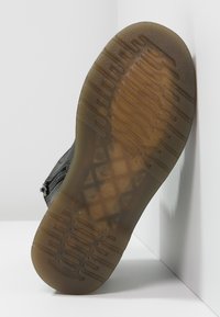 Bota negra con suela de goma texturizada, que presenta un patrón de dibujo en zigzag y un acabado ligeramente translúcido. Detalle de cremallera lateral visible.