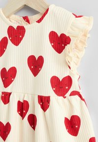 Geribbelde crèmekleurige jurk met een rood hartpatroon met glimlachende gezichten. Rimpelachtige mouwen en geplooide rokdetails, geschikt voor casual wear.