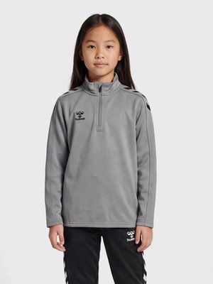 Joven chica de cabello largo y negro, de pie, vestida con una sudadera gris con cremallera hasta un cuarto y pantalones deportivos negros con logotipos y rayas.