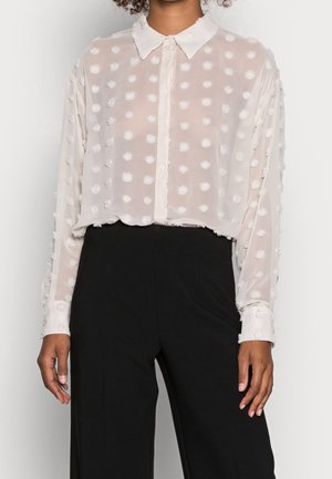 Ze draagt een doorzichtige witte blouse met opstaande polkadots en knoopmanchetten, gecombineerd met hoge zwarte broeken.