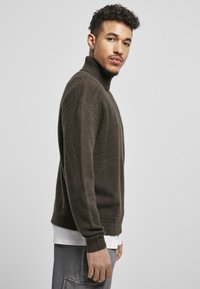 Mørkegrøn, tekstureret højhalset sweater med ribbede manchetter, afslappet pasform, båret over en hvid skjorte og stylet med grå bukser.