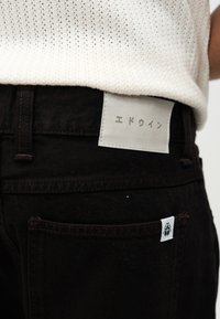 Black denim jeans with a white rectangular patch displaying Japanese characters and a small white tag on the back pocket.