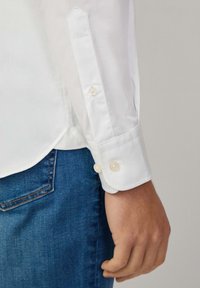 Chemise blanche à manches longues avec des poignets à boutons, présentant une texture lisse et un design ajusté, associée à un denim bleu.