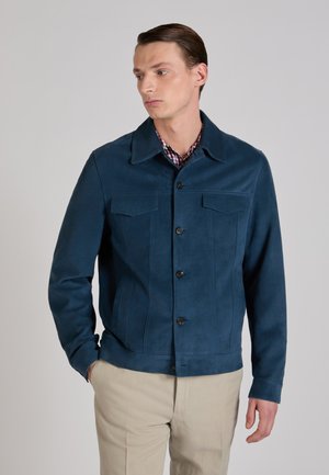 Man draagt een donkerblauwe knoopjasje over een geruit overhemd en beige broek, met één hand in de zak, kijkt naar zijn linkerzijde.