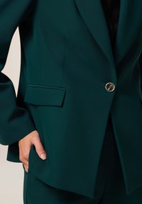 Blazer entallado en verde azulado con un solo botón de bronce, solapas con muesca y bolsillos laterales. Textura suave y corte estructurado.