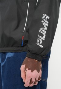 Jachetă neagră tip windbreaker cu mansete elastice, decorată cu logo-ul alb "PUMA" pe mânecă și accente roșii și albastre la tiv.