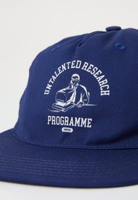 Tumedat sinine müts tekstureeritud kangast, millel on valged broderitud sõnad: "UNTALENTED RESEARCH PROGRAMME" ja graafika inimesest arvuti taga.