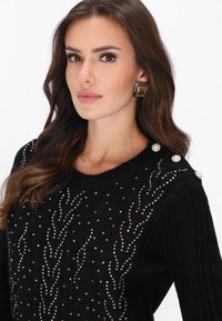 Zwarte gebreide trui met kleurrijke strasstenen versieringen in een golvend patroon, met parelaccenten op de schouders.