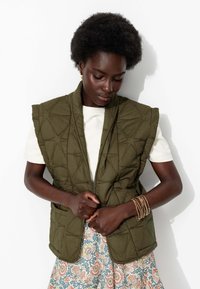 Gilet matelassé vert olive avec un col haut, présentant une couture géométrique. Porté par-dessus une chemise blanche, complété par une jupe à motifs.