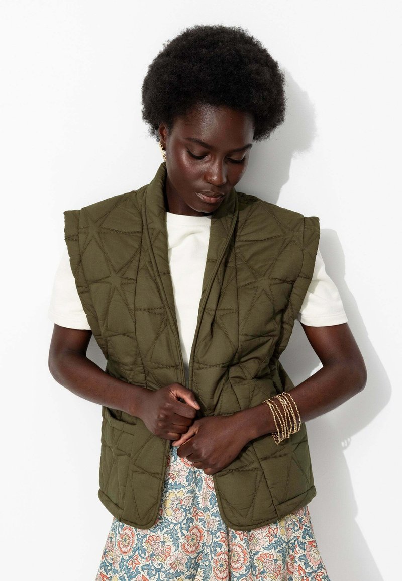 Gilet matelassé vert olive avec un col haut, présentant une couture géométrique. Porté par-dessus une chemise blanche, complété par une jupe à motifs.
