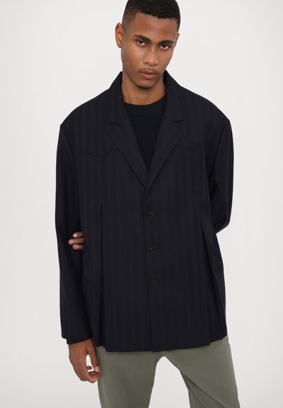 Marineblau gestreifter Oversized-Blazer mit Reverskragen, drei Knöpfen und Seitentaschen, über einem strukturierten schwarzen Stricktop getragen.