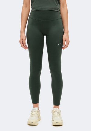 Una modelo lleva unos leggings Nike de color verde oscuro y un top a juego, con zapatillas deportivas de color crema, sobre un fondo blanco liso.