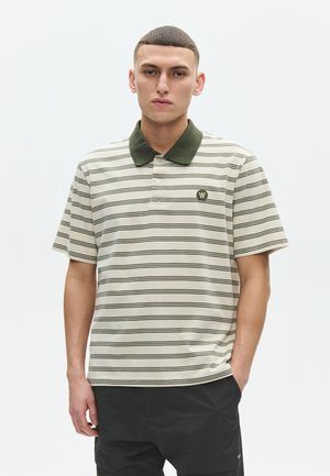 Unge mand med kort hår iført en creme- og sortstribet poloshirt med grøn krave og sorte bukser mod en ensfarvet hvid baggrund.