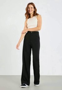Calças largas pretas com cintura alta, feitas de tecido suave. Combinadas com uma blusa creme sem mangas e ténis azul-marinho.
