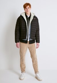 Chaqueta de abrigo negra con capucha forrada de piel sintética blanca, superpuesta a una camisa de mezclilla, combinada con pantalones de pana color beige y zapatillas blancas.
