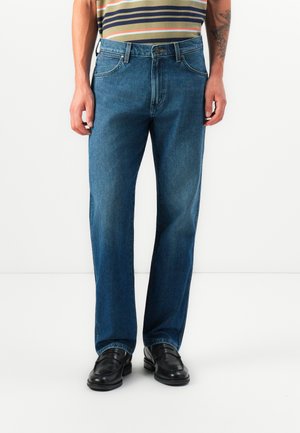 Hombre que lleva vaqueros ajustados de color azul, mocasines de cuero negros y una camisa a rayas con los brazos relajados a los lados.