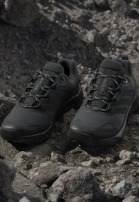 Coppia di scarpe da trail nere da esterno con lacci riflettenti che riposano su terreno roccioso.