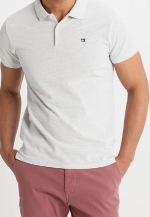 Hombre con una camisa polo de manga corta de color gris claro con un pequeño logo en el pecho y pantalones malva, manos en los bolsillos.