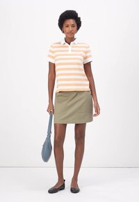 Polo-shirt met korte mouwen in oranje en witte strepen, gecombineerd met een kaki rok. Model houdt een blauwe tas vast. Platte zwarte schoenen.