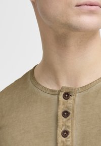 Maglietta henley verde oliva a maniche corte con scollatura arrotondata e tre bottoni marroni sul davanti. Tessuto morbido e texturizzato.