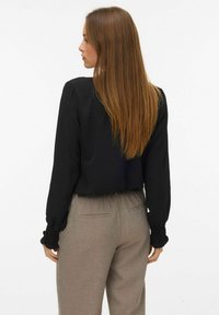 Sieviete ar gariem taisniem matiem, tērpusies melnā garā piedurkņu blūzē un bēšās biksēs, stāv apejo uz vienkārša fona.