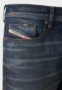Blue denim jeans featuring a slightly faded wash, orange stitching, and a branded label in the back pocket with white and red details.