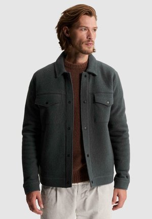 Homme aux cheveux châtain clair portant une veste boutonnée vert foncé par-dessus un pull marron et un pantalon clair, regardant vers la droite.