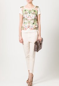 Blomstermønstrede top med en cremefarvet bund, parret med lyse bukser. Modellen holder en brun taske. Skoene er beige højhælede. Enkel design, afslappet pasform.