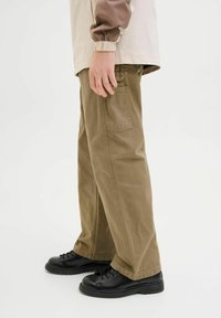 Pantaloni beige a gamba larga con una finitura testurizzata e una tasca posteriore, abbinati a scarpe nere con lacci. Mani parzialmente visibili di lato.