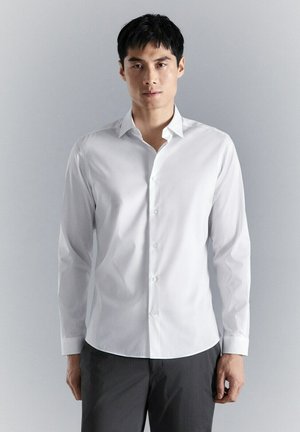 Junger Mann steht, trägt ein weißes Hemd mit Knöpfen und dunkle Hose, vor einem schlichten hellgrauen Hintergrund.