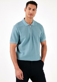 Gaiši zils polo krekls ar īsām piedurknēm, pus-izvelkams priekšpuse, teksturēta auduma un klasiskā apkakle. Apvienots ar tumšiem biksēm.